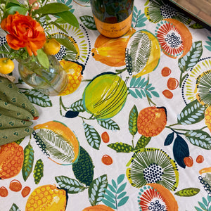 Close up of table top covered in Agrume Fruits tablecloth. The tablecloth background is white and the pattern  features lemons, limes, oranges, kiwis and leaves. Vibrants colors of orange, yellow and green.