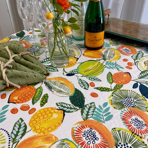 Tabletop with Agrume Fruits tablecloth, champagne bottle, floral arrangement and coordinating green napkins. Tablecloth has white background with lemons, limes, oranges and kiwi pattern.
