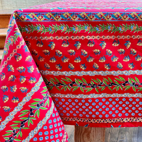 Colorful patterned tablecloth with a red background and stripes of olive sprigs, floral designs, vines, and french medallions on a wooden surface.