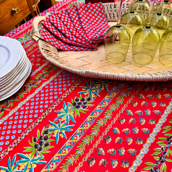 Bright red tablecloth with  pattern of olive sprigs, flowers and french medallions. A wicker tray on top of tablecloth holding red and blue matching napkins and green wine glasses.
