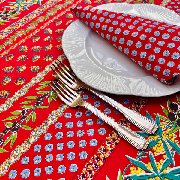 Decorative table setting with floral-patterned tablecloth. Pattern is stripes of olives, leaves, mimosa flowers and medallions in turquoise, green, blue, black and yellow on a vibrant red background. While plate, silver forks, coordinating red and blue napkin